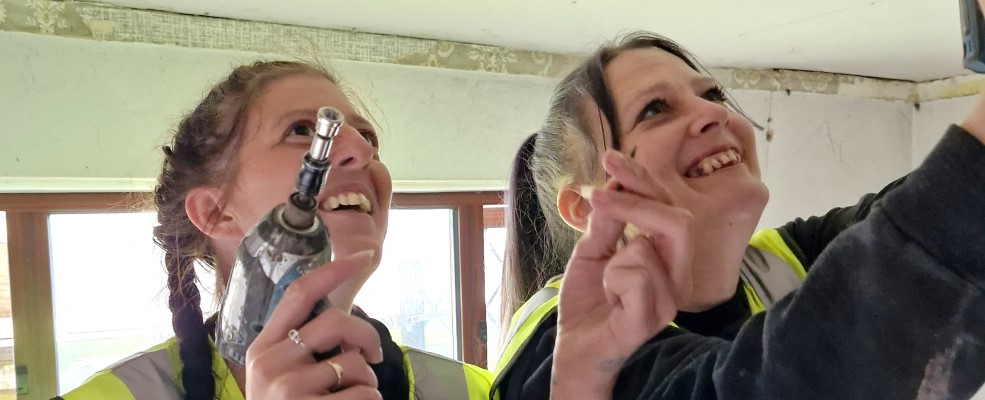 Close-up of two women working on a construction site, one is holding a drill. They are both smiling.