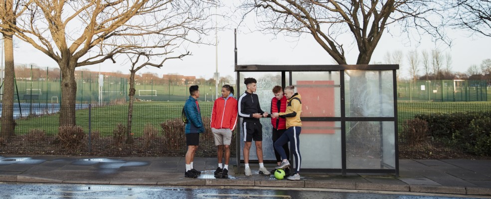 Wide shot of five young men stand in a line at the bus stop after playing five a side football.