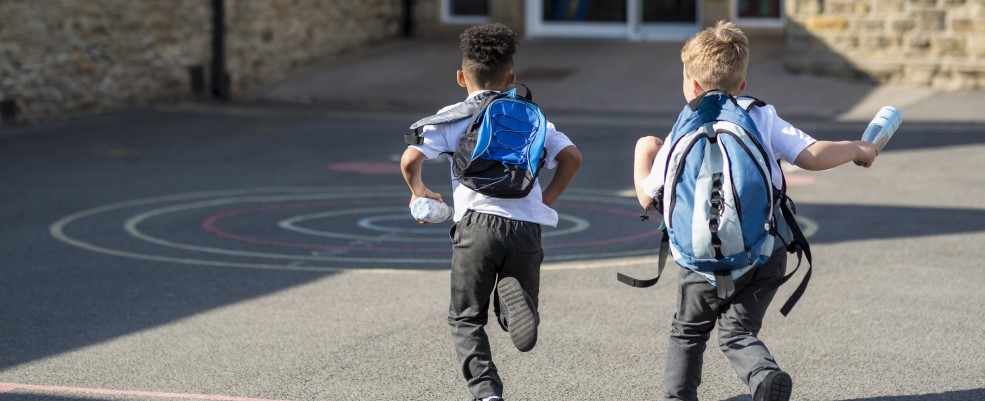 Rear view of two boys running in their school yard. They are running towards the door with their backpacks on.
