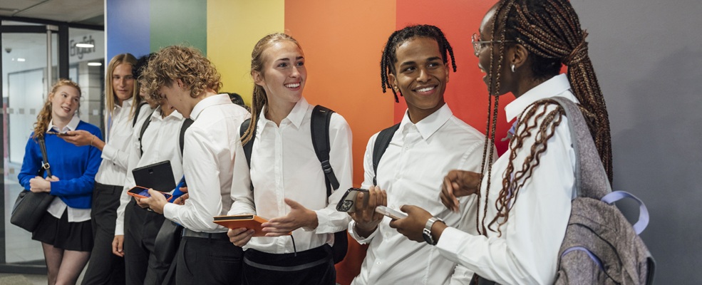 Students wearing school uniform, standing together in the lobby of a school in Gateshead, North East England. They are waiting to go to class and are bonding and laughing with each other. Some student are using their mobile phone.